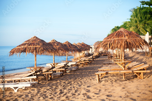 Rows of empty bamboo chaise lounge and thatched umbrellas on lonely white sand beach  on the blue sea and green palm trees background  nobody  design tourist poster  travel banner  relax vacation card