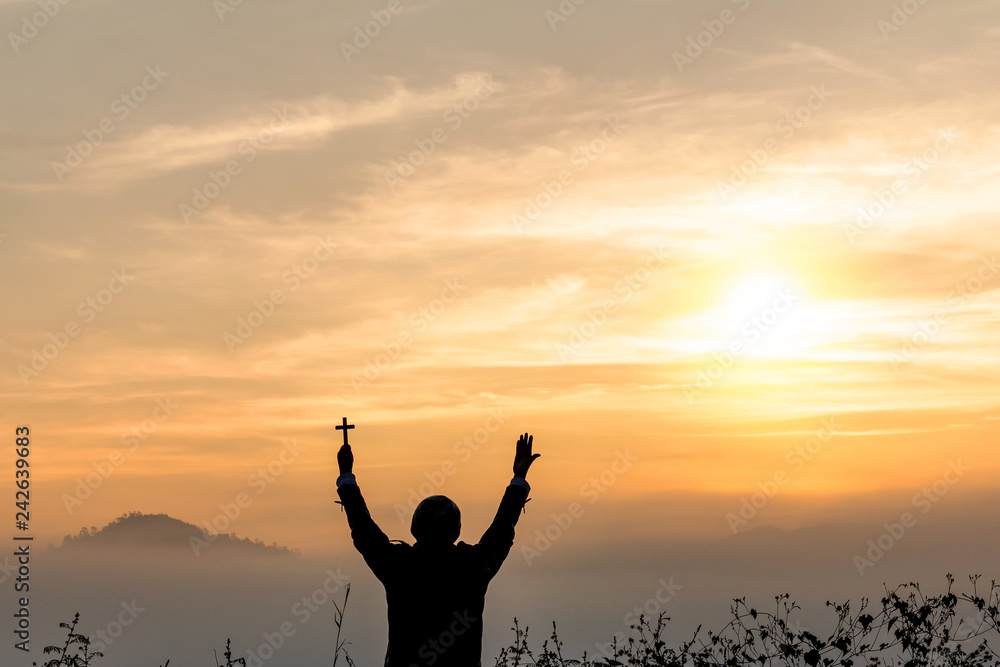 Silhouette of human praying to the GOD while holding a crucifix symbol with bright sunbeam on the sky