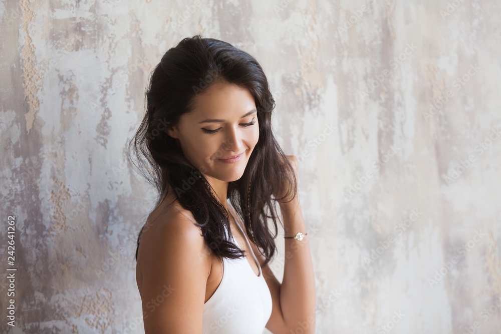 Charming young brunette woman shyly smiles and looks down a laser against a background of a concrete wall Charming young brunette woman shyly smiles and looks down a laser against a background of a concrete wall