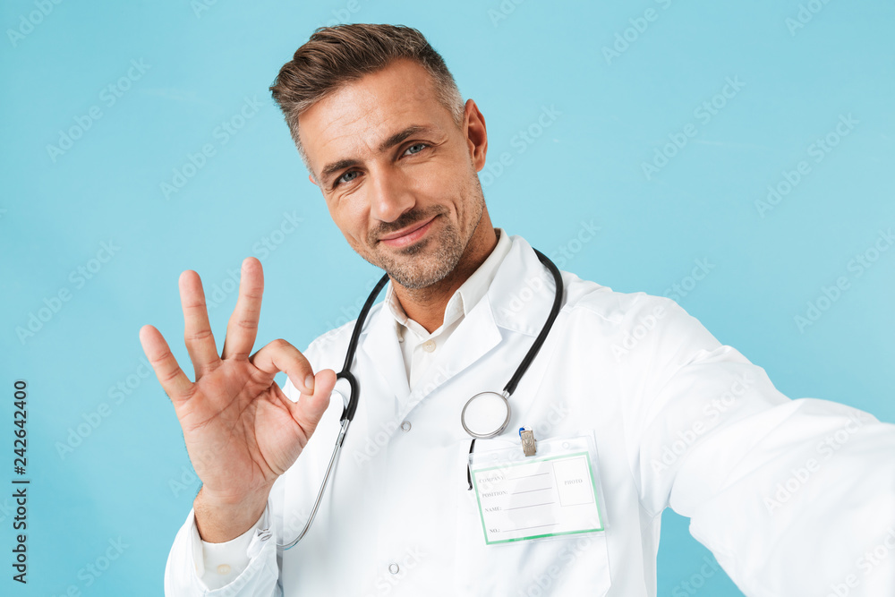 Portrait of positive medical doctor with stethoscope taking selfie photo, while standing isolated over blue background