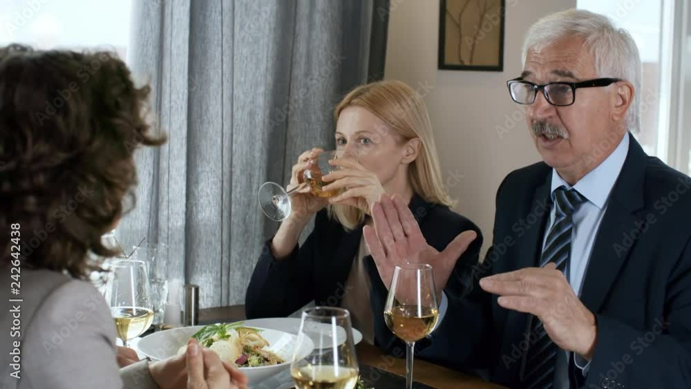 Senior entrepreneur and his female colleague sitting together at table with wine and food on it and speaking with business partners while having dinner in the restaurant Senior entrepreneur and his female colleague sitting together at table with wine and food on it and speaking with business partners while having dinner in the restaurant