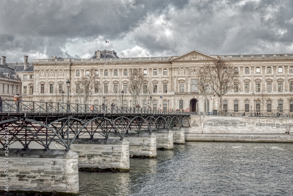 People walking on Pont des Arts in winter with the Louvre Museum in background - Paris, France