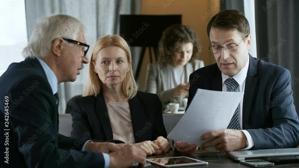 Senior entrepreneur and middle aged businessman discussing document while sitting with female colleague at restaurant table during break Senior entrepreneur and middle aged businessman discussing document while sitting with female colleague at restaurant table during break