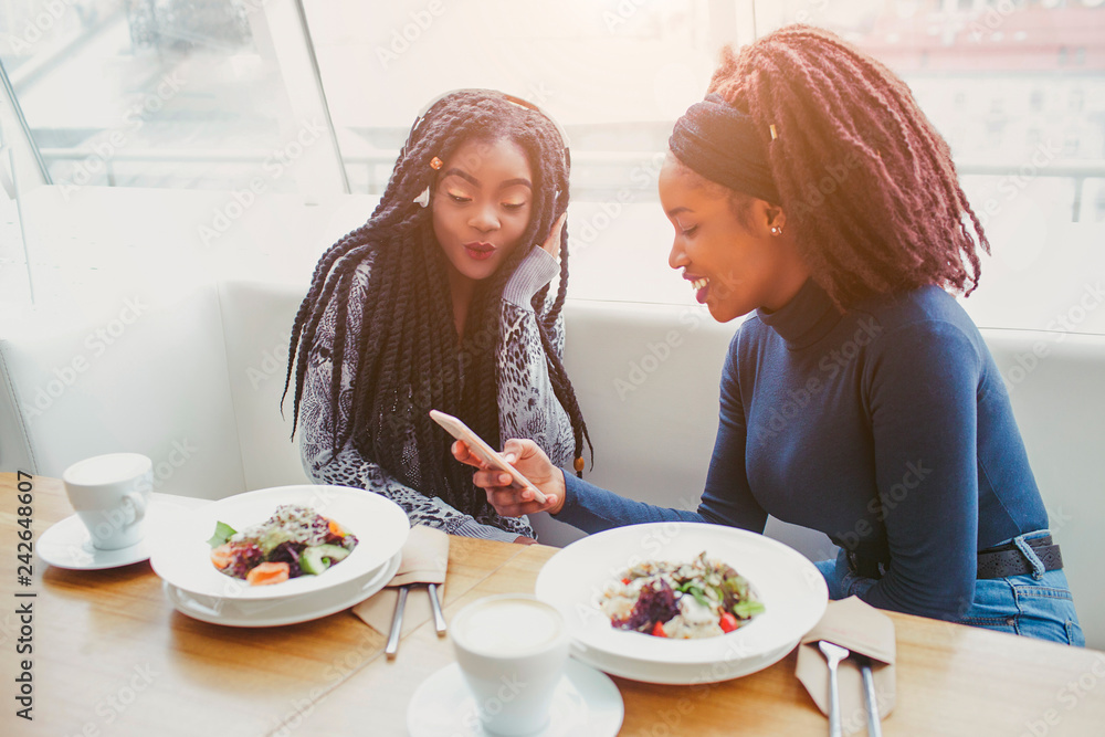 Two african american women sit in cafe at table. They look at phone. Food in balls are on table. Two african american women sit in cafe at table. They look at phone. Food in balls are on table.