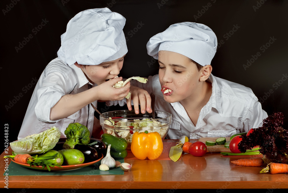 The boys in the cooks costumes cut the salad in the kitchen. Children prepare healthy food. Friendly cooperation of cooks. Isolated on black background. The boys in the cooks costumes cut the salad in the kitchen. Children prepare healthy food. Friendly cooperation of cooks. Isolated on black background.