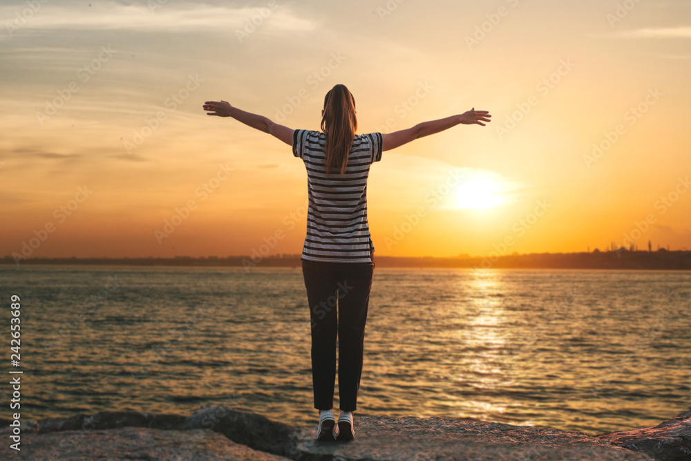 Fototapeta premium A girl in solitude stands on the shores of the Bosphorus in Istanbul in Turkey and raises her hands up showing how she is free and how she likes. There is a beautiful sunset ahead.