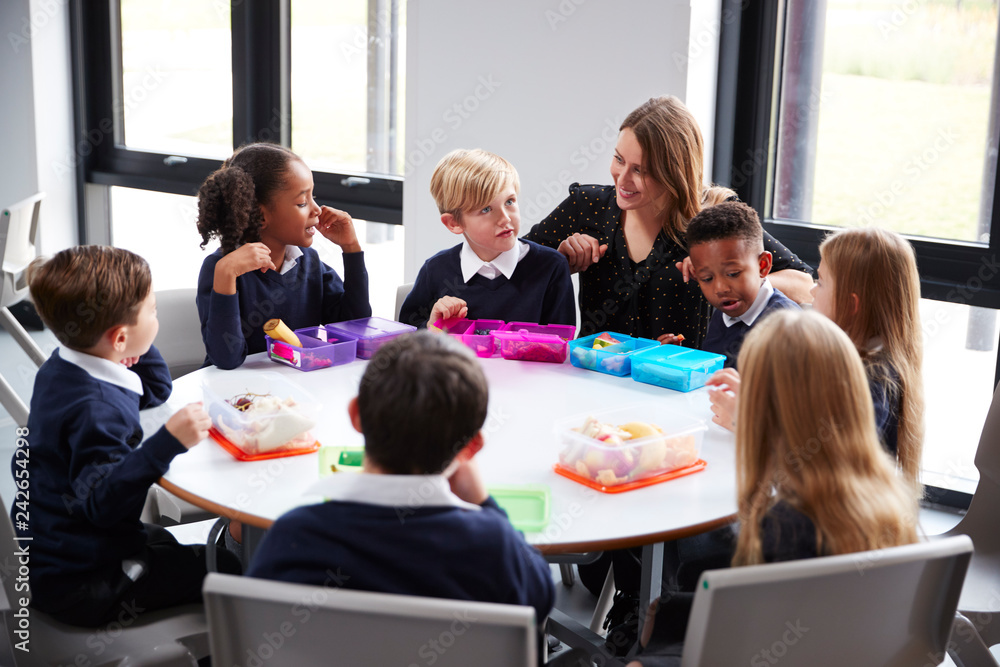 Female teacher kneeling to talk to a group of primary school kids sitting together at a round table eating their packed lunches