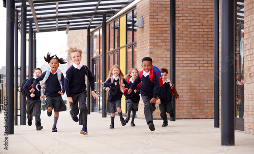 Primary school kids, wearing school uniforms and backpacks, running on a walkway outside their school building, front view photo
