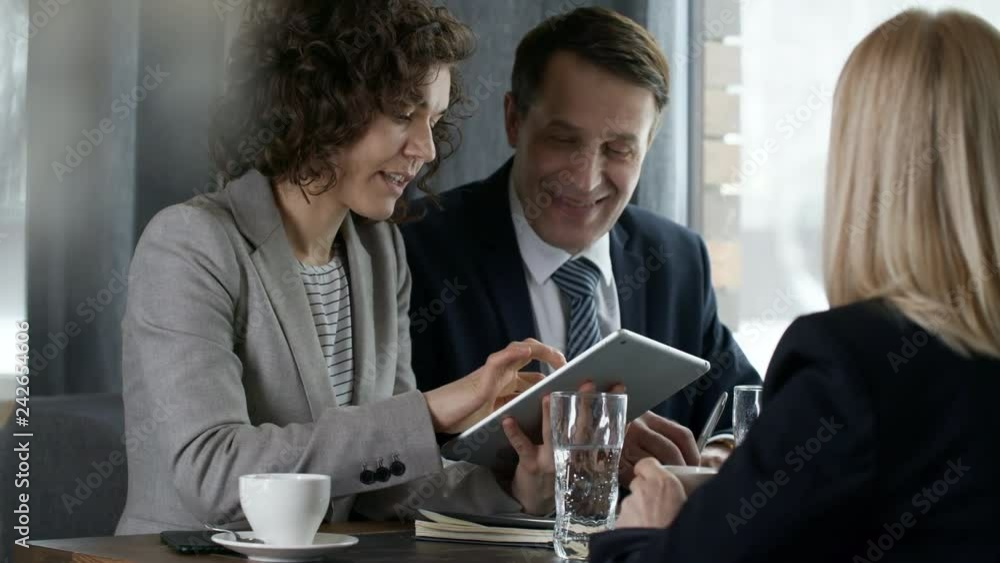Experienced middle aged businessman wearing formal suit and glasses sitting at restaurant table with young female colleague and explaining something to partners during lunch Experienced middle aged businessman wearing formal suit and glasses sitting at restaurant table with young female colleague and explaining something to partners during lunch