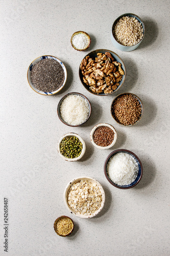 Variety of raw uncooked grains superfood cereal chia, linen, sesame, mung bean, walnuts, tapioca, wheat, buckwheat, oatmeal, coconut, rice in ceramic bowls grey spotted background. Flat lay space