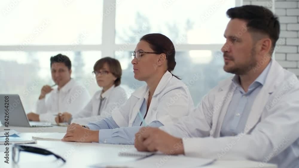 Professional female practitioners in lab coats sitting at desk with male colleagues and asking questions and discussing something with teacher during health seminar Professional female practitioners in lab coats sitting at desk with male colleagues and asking questions and discussing something with teacher during health seminar