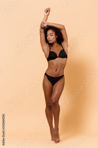 Full length portrait of pretty woman wearing black lingerie, standing isolated over beige background