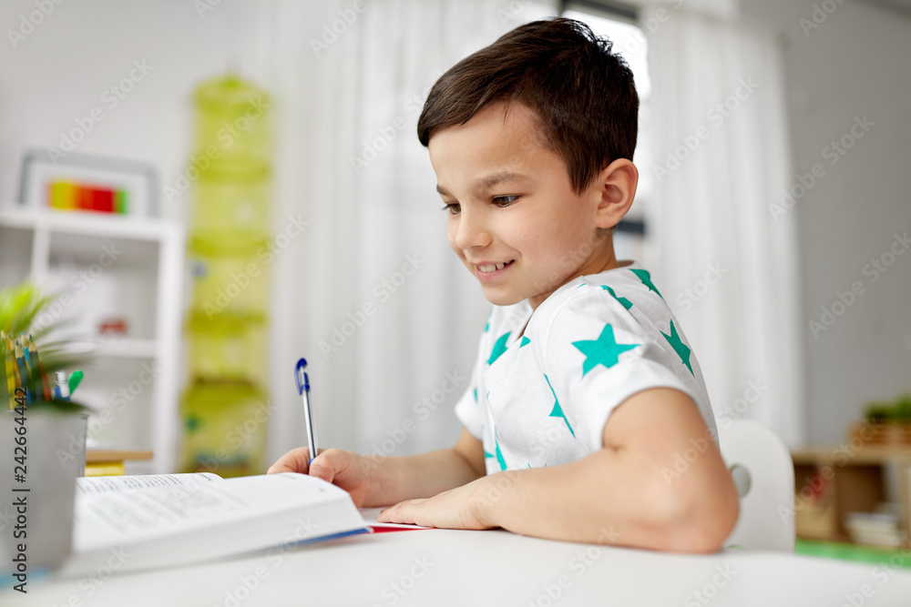 education, childhood and people concept - happy student boy with book writing to notebook at home education, childhood and people concept - happy student boy with book writing to notebook at home