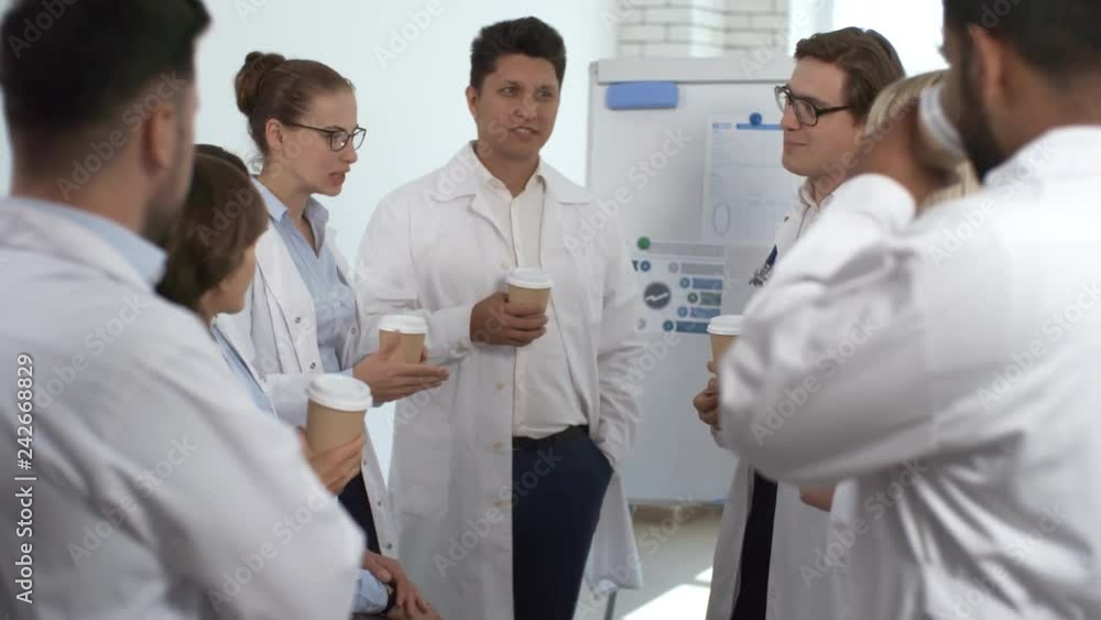 Group of multiethnic healthcare workers in lab coats drinking coffee, smiling and discussing presentation on flipchart after meeting Group of multiethnic healthcare workers in lab coats drinking coffee, smiling and discussing presentation on flipchart after meeting