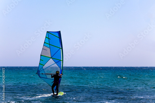Rider in Wetsuit and Helmet Balancing on a Surfboard with Sail Mast and Bar. Clear Sky and Blue Wave. Surfer Trying out the Outdoor Water Sport Adventure © Artur
