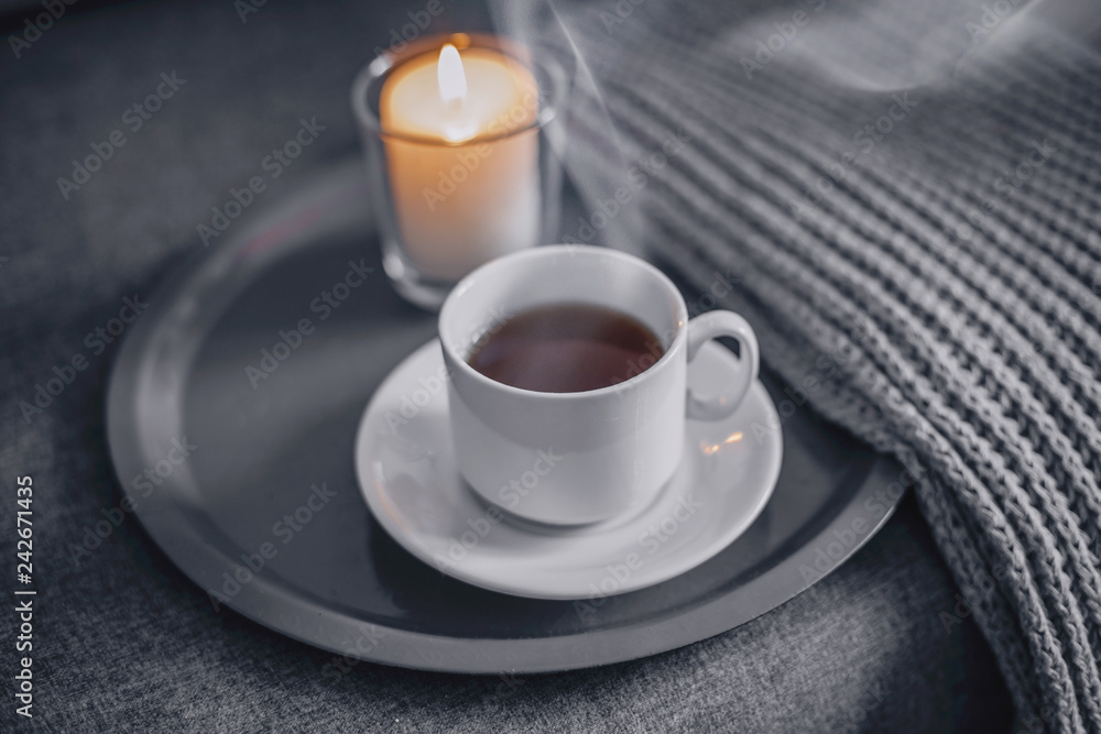 Cup of hot coffee with a candle on a gray tray, next to a knitted gray scarf and a white sweater. Concept cozy winter home relax Cup of hot coffee with a candle on a gray tray, next to a knitted gray scarf and a white sweater. Concept cozy winter home relax