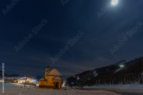 Night view of a small church at the San Pellegrino Pass in winter, with the snow-covered ski slopes in the background, Dolomites, Italy