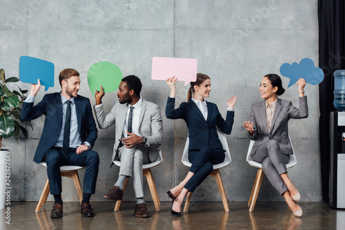 happy multiethnic businesspeople holding speech bubbles and thought bubble while sitting in waiting hall