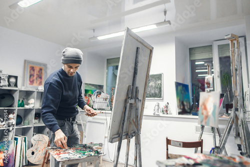 Young, positive artist works, paints a picture of oil paints in a cozy studio. Artist with a canvas on an easel places an oil paint on a palette. Process of creating a picture. Copyspace