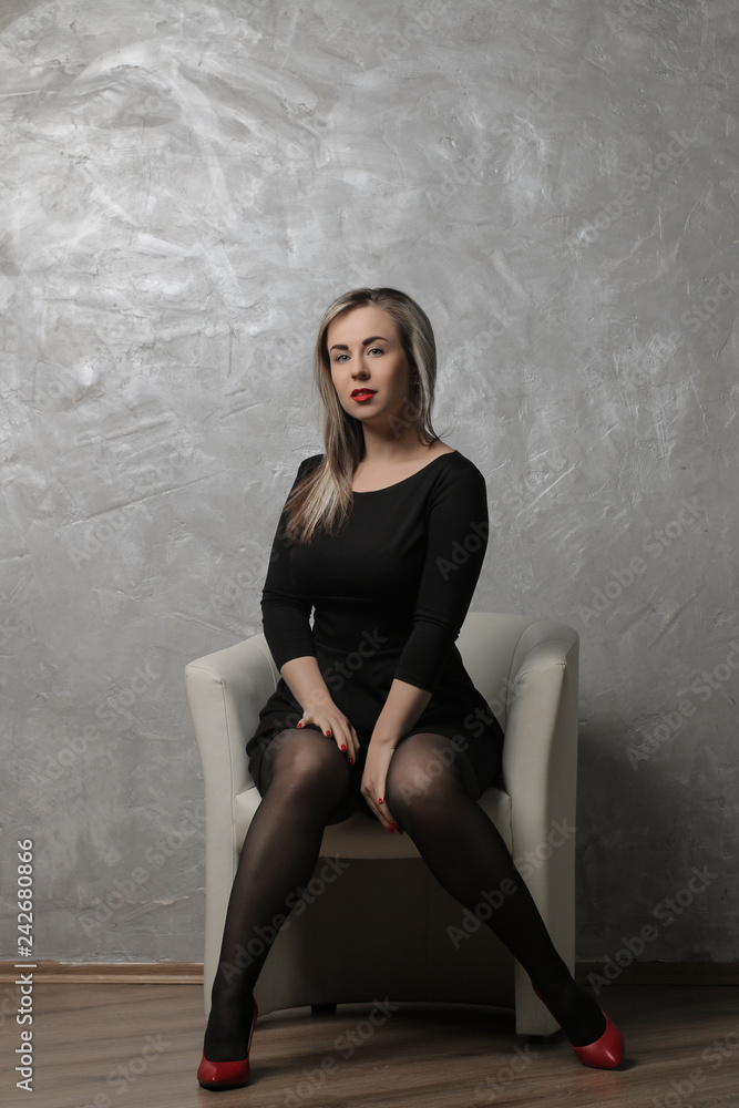 Fototapeta premium Beautiful girl with red lips and in a black dress sitting in a chair against the background of a gray concrete wall. emotion. different poses