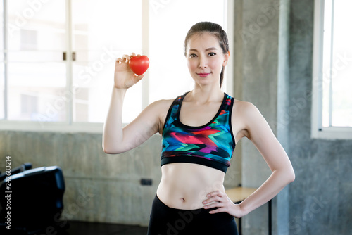 Beautiful white Asian woman wearing sportswear smile sitting on hand holding red heart in gym. Lose weight by exercising regularly. Control the amount of food useful. Strong body and beautiful skin.