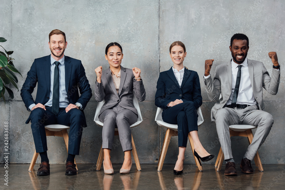 smiling multiethnic businesspeople sitting on chairs and cheering with clenched fists in waiting hall smiling multiethnic businesspeople sitting on chairs and cheering with clenched fists in waiting hall