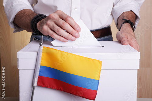 A Colombian citizen inserting a ballot into a ballot box. Colombia flag in front of it