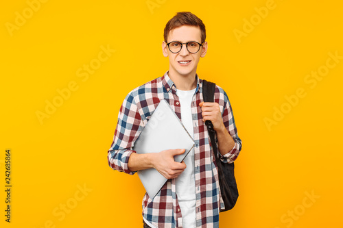Happy guy with glasses, a man in a plaid shirt, with a closed laptop in hand, on a yellow background