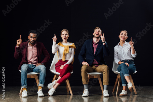 multiethnic group of people sitting on chairs and showing idea gestures isolated on black