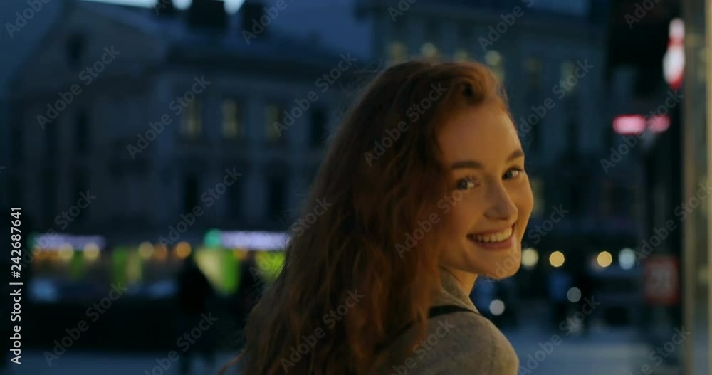 Close up of the red haired young cheerful Caucasian girl walking the street in the evening and smiling to the camera. Rear. Back view. Close up of the red haired young cheerful Caucasian girl walking the street in the evening and smiling to the camera. Rear. Back view.