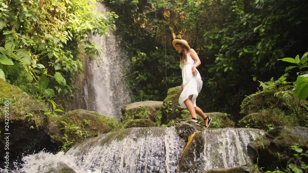 Attractive Girl in White Dress and Straw Hat Walking Barefoot near Small Waterfall in Tropical Rainforest Jungle. Carefree Lifestyle Travel 4K Slowmotion Footage. Bali, Indonesia. Attractive Girl in White Dress and Straw Hat Walking Barefoot near Small Waterfall in Tropical Rainforest Jungle. Carefree Lifestyle Travel 4K Slowmotion Footage. Bali, Indonesia.