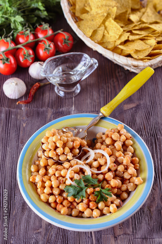 Boiled chickpeas, seasoned with vegetable oil, ground red pepper, sesame seeds and onion rings in a bowl with old spoon on a wooden background next to garlic and tomatos and corn chips, vertical orien photo