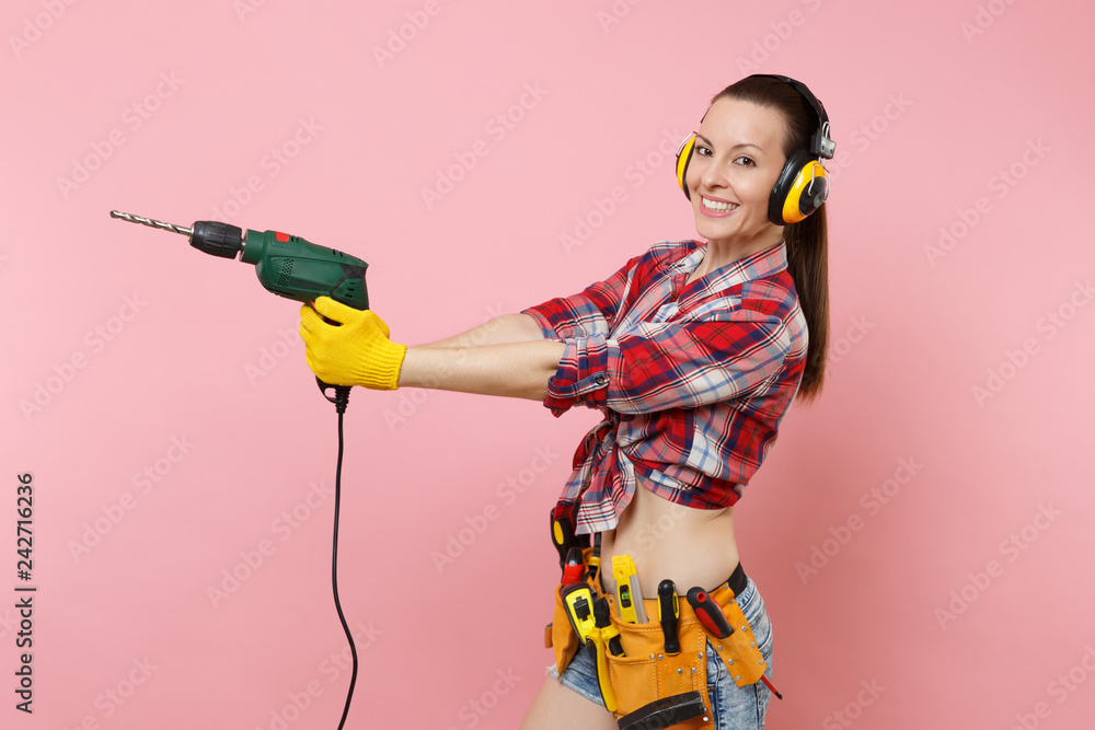 Energy sexy handyman woman in gloves, noise insulated headphones, kit tools belt full of instruments holding power electric drill isolated on pink background. Female in male work. Renovation concept Energy sexy handyman woman in gloves, noise insulated headphones, kit tools belt full of instruments holding power electric drill isolated on pink background. Female in male work. Renovation concept