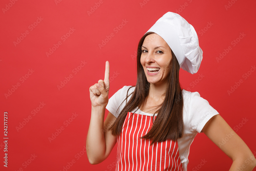Housewife female chef cook or baker in red striped apron, white t-shirt, toque chefs hat isolated on red wall background. Close up portrait of housekeeper brunette woman. Mock up copy space concept. Housewife female chef cook or baker in red striped apron, white t-shirt, toque chefs hat isolated on red wall background. Close up portrait of housekeeper brunette woman. Mock up copy space concept.
