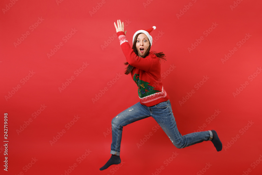 Amazed Santa girl in Christmas hat jumping, rising hands spreading legs, keeping mouth wide open isolated on red background. Happy New Year 2019 celebration holiday party concept. Mock up copy space.