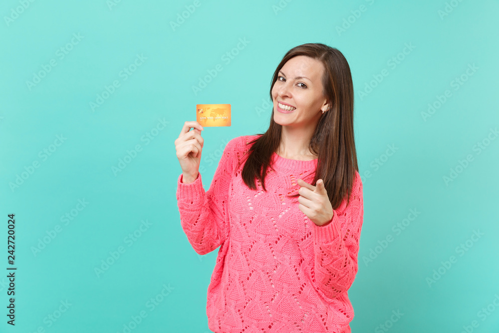 Smiling young woman in knitted pink sweater pointing index finger on camera, holding in hand credit card isolated on blue wall background studio portrait. People lifestyle concept. Mock up copy space. Smiling young woman in knitted pink sweater pointing index finger on camera, holding in hand credit card isolated on blue wall background studio portrait. People lifestyle concept. Mock up copy space.