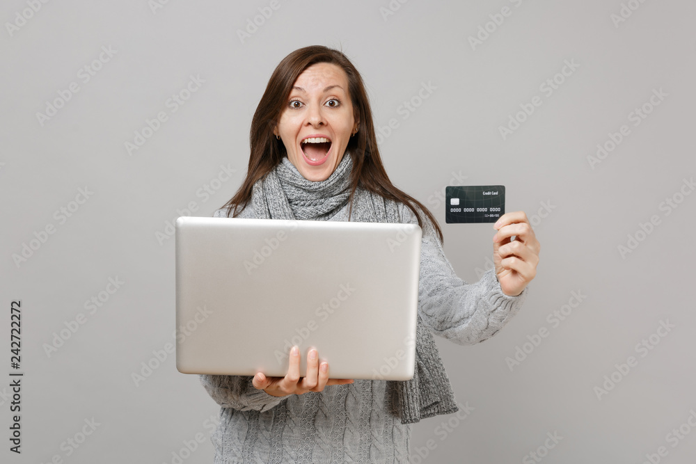 Surprised woman in sweater, scarf with opened mouth working on laptop pc computer, hold credit bank card isolated on grey background. Healthy lifestyle online treatment consulting cold season concept. Surprised woman in sweater, scarf with opened mouth working on laptop pc computer, hold credit bank card isolated on grey background. Healthy lifestyle online treatment consulting cold season concept.