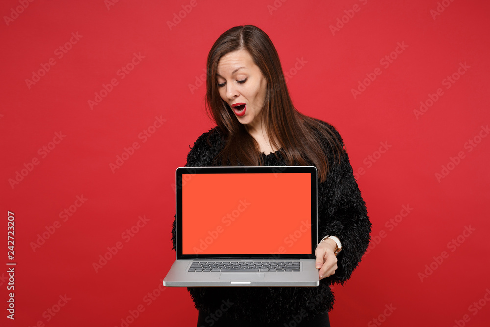 Naklejka premium Bewildered young woman in black fur sweater looking on laptop pc computer with blank empty screen isolated on bright red wall background. People sincere emotions lifestyle concept. Mock up copy space.