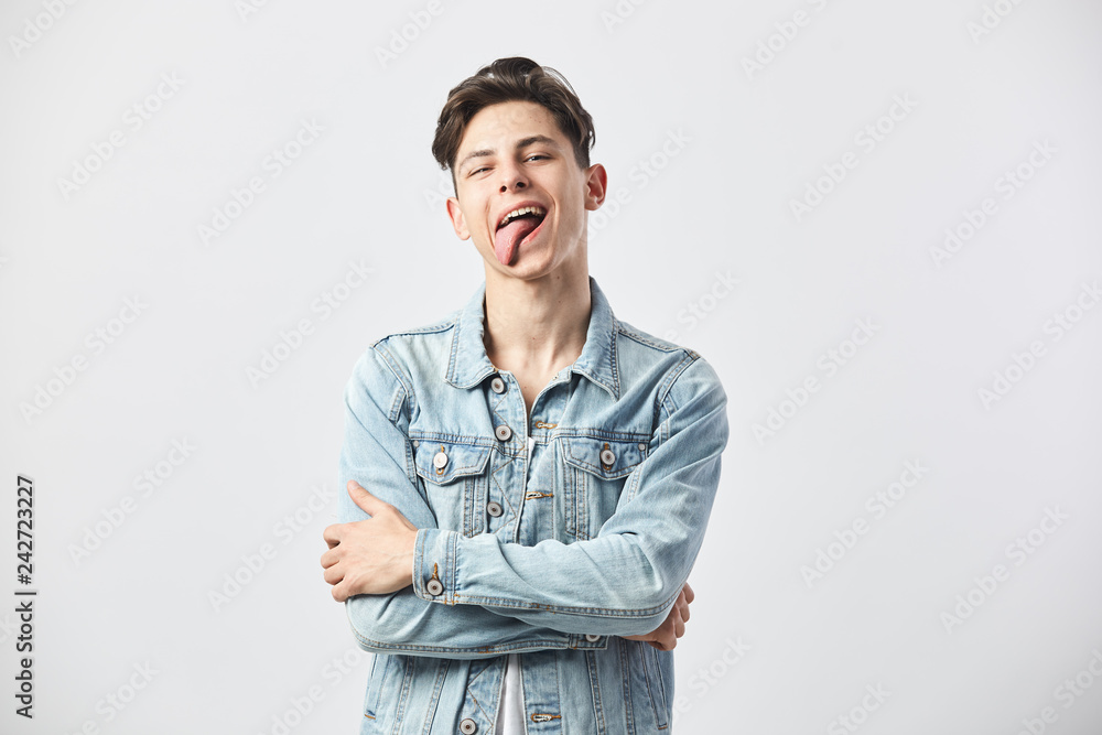 Funny dark-haired guy dressed in a white t-shirt and a denim jacket shows his tongue on the white background in the studio Funny dark-haired guy dressed in a white t-shirt and a denim jacket shows his tongue on the white background in the studio