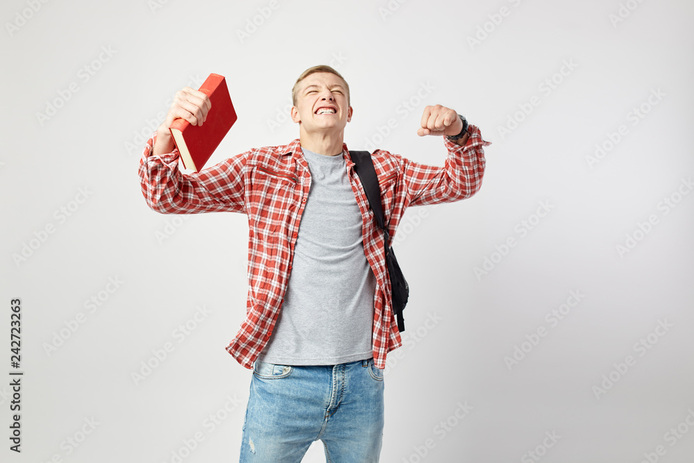 Fototapeta premium Joyful blond guy with black backpack on his shoulder dressed in a white t-shirt, red checkered shirt and jeans holds book in his hand on the white background in the studio
