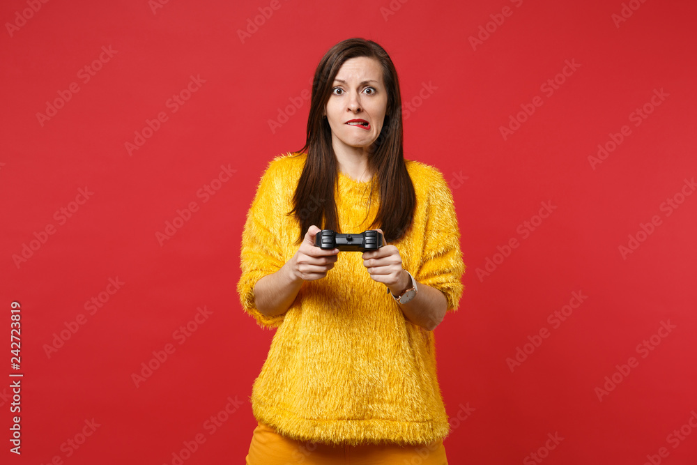 Portrait of concerned young woman in yellow fur sweater biting lips, playing video game with joystick isolated on bright red background. People sincere emotions, lifestyle concept. Mock up copy space. Portrait of concerned young woman in yellow fur sweater biting lips, playing video game with joystick isolated on bright red background. People sincere emotions, lifestyle concept. Mock up copy space.