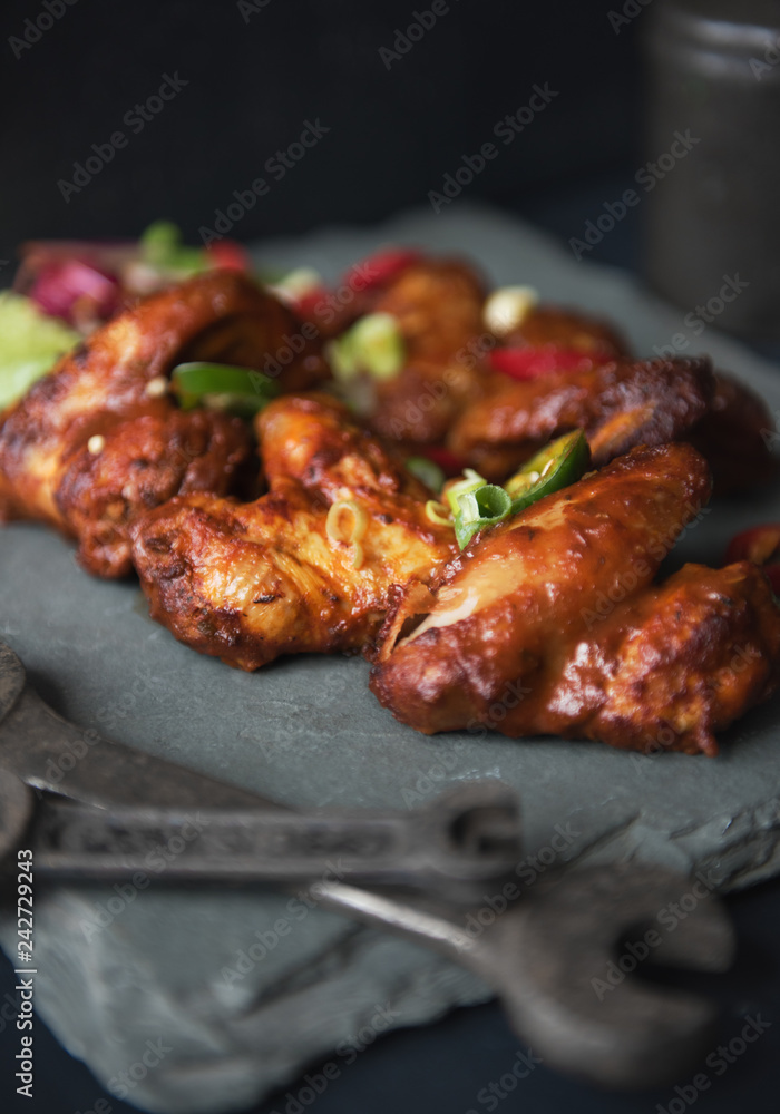 A beautiful platter slate plate full of organic delicious golden and crispy bbq chicken wings, served in a dark photography mechanic style environment. Surrounded by mechanic tools. food fashion.