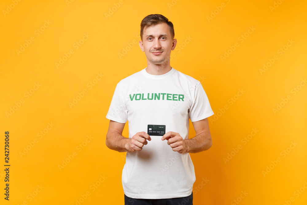 Portrait of man in white t-shirt with written inscription green title volunteer hold in hand credit bank card isolated on yellow background. Voluntary free assistance help, charity grace work concept. Portrait of man in white t-shirt with written inscription green title volunteer hold in hand credit bank card isolated on yellow background. Voluntary free assistance help, charity grace work concept.