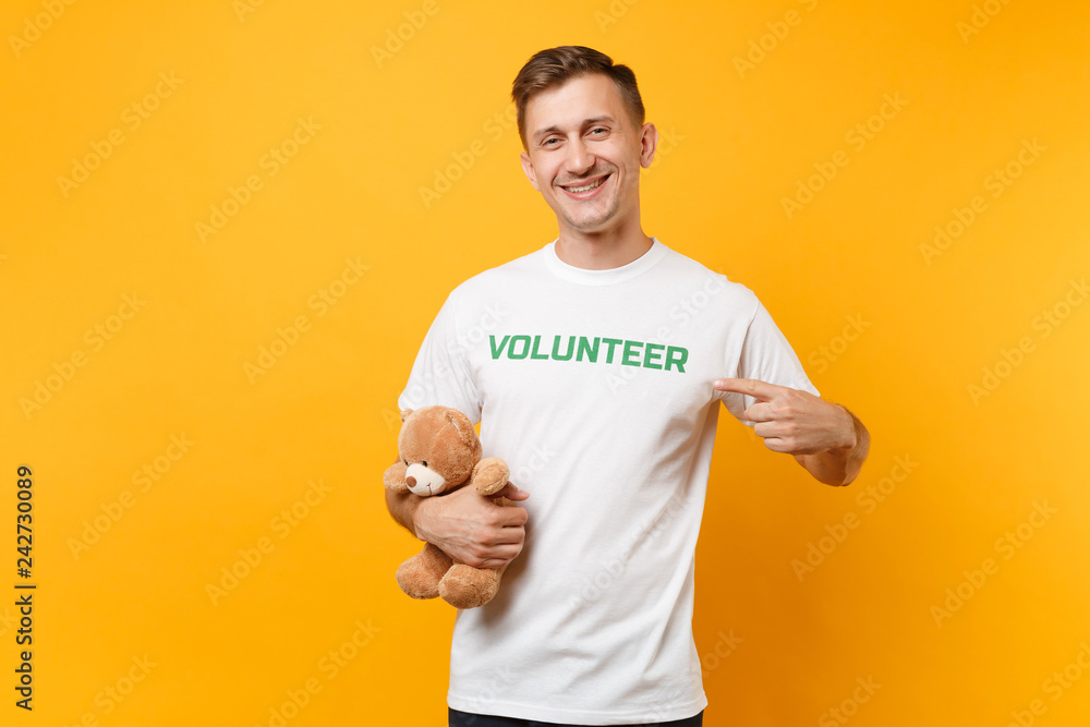 Portrait of good man in white t-shirt with written inscription green title volunteer hold teddy bear plush toy isolated on yellow background. Voluntary free assistance help, charity grace work concept Portrait of good man in white t-shirt with written inscription green title volunteer hold teddy bear plush toy isolated on yellow background. Voluntary free assistance help, charity grace work concept
