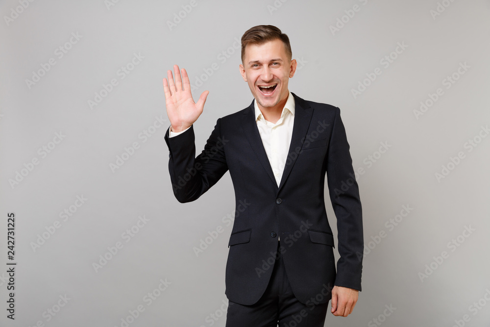 Cheerful young business man in classic black suit, shirt waving and greeting with hand as notices someone isolated on grey background. Achievement career wealth business concept. Mock up copy space. Cheerful young business man in classic black suit, shirt waving and greeting with hand as notices someone isolated on grey background. Achievement career wealth business concept. Mock up copy space.