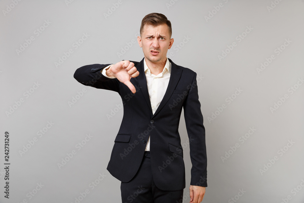 Portrait of dissatisfied young business man in classic black suit, shirt showing thumb down isolated on grey wall background in studio. Achievement career wealth business concept. Mock up copy space. Portrait of dissatisfied young business man in classic black suit, shirt showing thumb down isolated on grey wall background in studio. Achievement career wealth business concept. Mock up copy space.