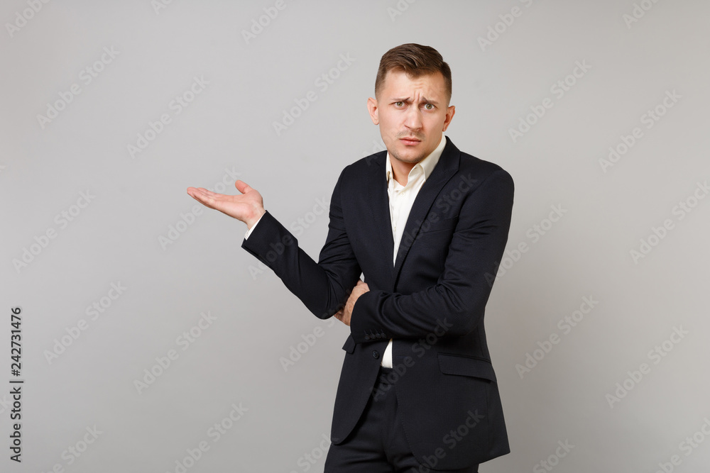 Puzzled young business man in classic black suit, white shirt spreading pointing hands aside isolated on grey wall background in studio. Achievement career wealth business concept. Mock up copy space. Puzzled young business man in classic black suit, white shirt spreading pointing hands aside isolated on grey wall background in studio. Achievement career wealth business concept. Mock up copy space.