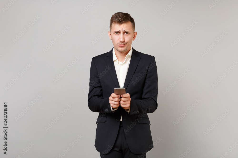 Puzzled young business man in classic black suit, shirt using mobile phone and typing sms message isolated on grey background in studio. Achievement career wealth business concept. Mock up copy space. Puzzled young business man in classic black suit, shirt using mobile phone and typing sms message isolated on grey background in studio. Achievement career wealth business concept. Mock up copy space.