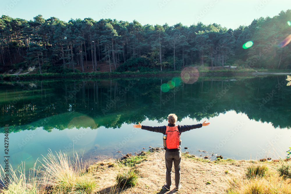 Back view young tourist man with backpack arms raised up to sky, celebrating freedom standing near forest lake. Exploring the world. Walking adventure. Selective focus, copy space. Blue poole, Uk. Back view young tourist man with backpack arms raised up to sky, celebrating freedom standing near forest lake. Exploring the world. Walking adventure. Selective focus, copy space. Blue poole, Uk.