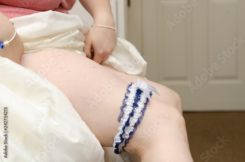 A blue and white lace Garter on the leg of a bride, A pale skinned sexy bride in a luxury wedding dress showing her silky garter. Woman have a final preparation for wedding ceremony